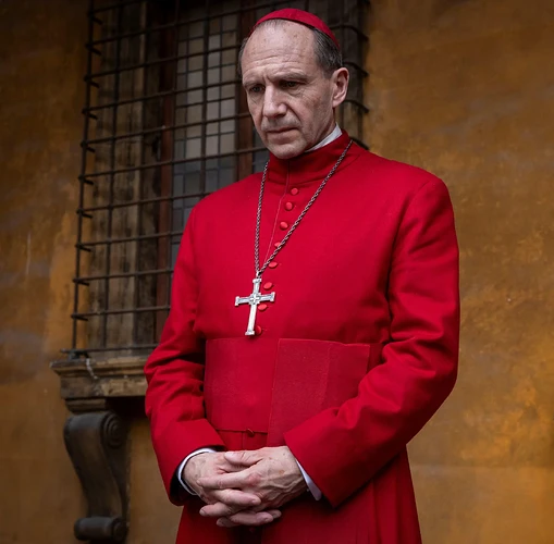 Ralph Fiennes as a cardinal in a red cassock with a cross, looking down with clasped hands.