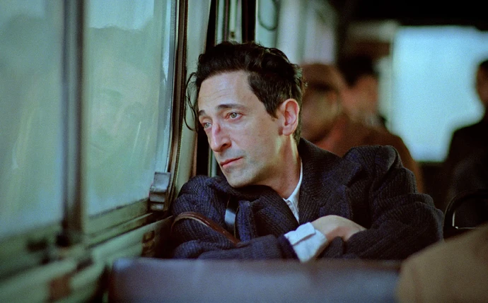Adrien Brody as an older man with tousled hair looking out a bus window.