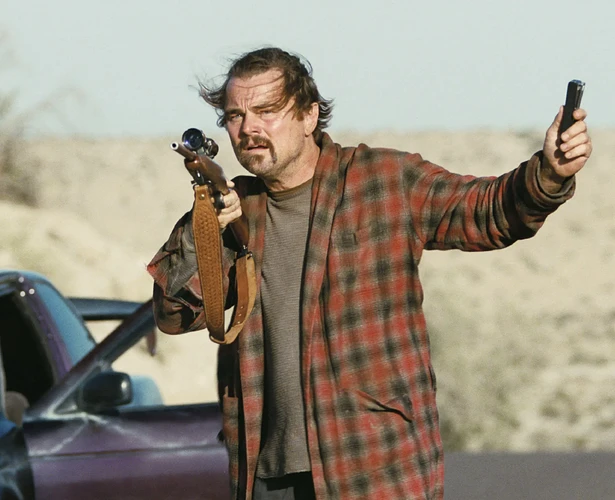 Leonardo DiCaprio holding a rifle and a cell phone, standing next to a car on an empty road.