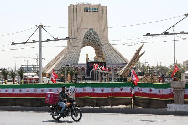 A person riding a motorcycle past a series of Iranian flags and missiles on display in front of a large monument in Tehran.