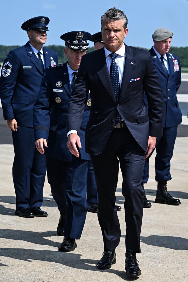 Pete Hegseth, wearing a dark suit and dark tie, walking on a tarmac next to other military officials.