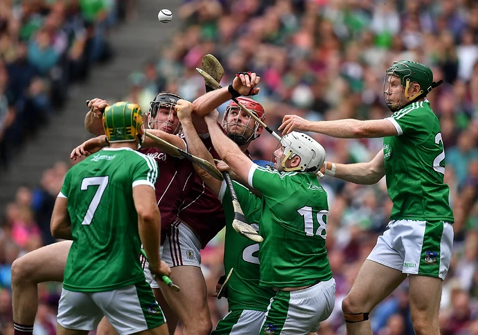 Six hurling players, five in green and white and one in maroon and white, converge as a white sliotar is in the air above them.