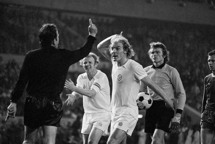 Football players Terry Yorath and Billy Bremner of Leeds United argue with referee Michel Kitabdjian during a game, while Bayern Munich goalkeeper Sepp Maier looks on.