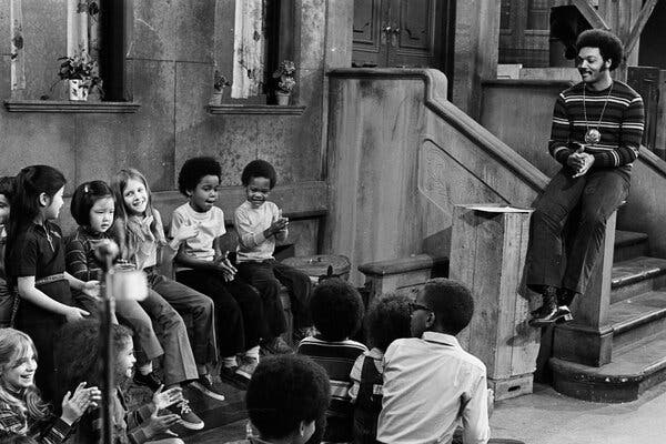 A black-and-white image of a man with an Afro and wearing a large necklace medallion, who sits on the stoop of the "Sesame Street" set, modeled to look like a New York City block. He is smiling and addressing a group of smiling, clapping children of different ages, genders and races.
